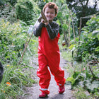 A child in red overalls & wellies walks along a path in a lush garden, carrying a gardening tool with a big smile.